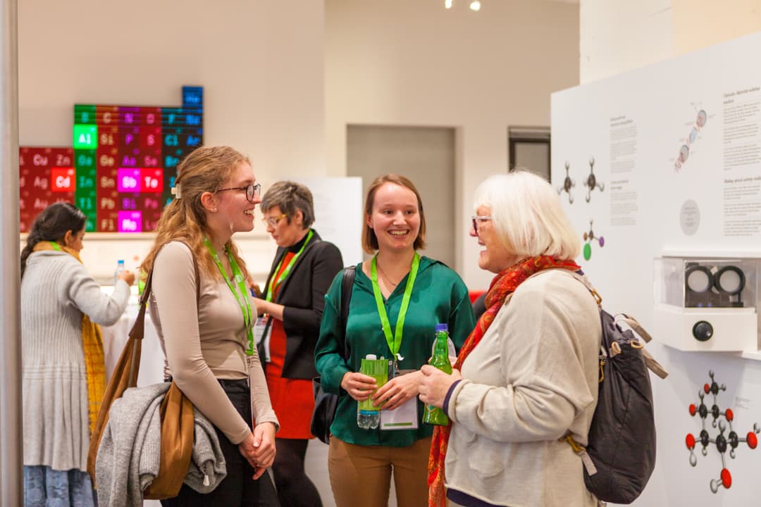 Mehrere Personen stehen in einem Ausstellungsraum und unterhalten sich. Im Hintergrund sind bunte Tafeln mit chemischen Elementsymbolen an der Wand angebracht. Auf weißen Tafeln sind Molekülmodelle und wissenschaftliche Grafiken zu sehen. Einige Personen tragen grüne Namensschilder um den Hals. Eine Person hält zwei grüne Flaschen in der Hand. Die Kleidung ist überwiegend leger, mit Schals und Jacken.