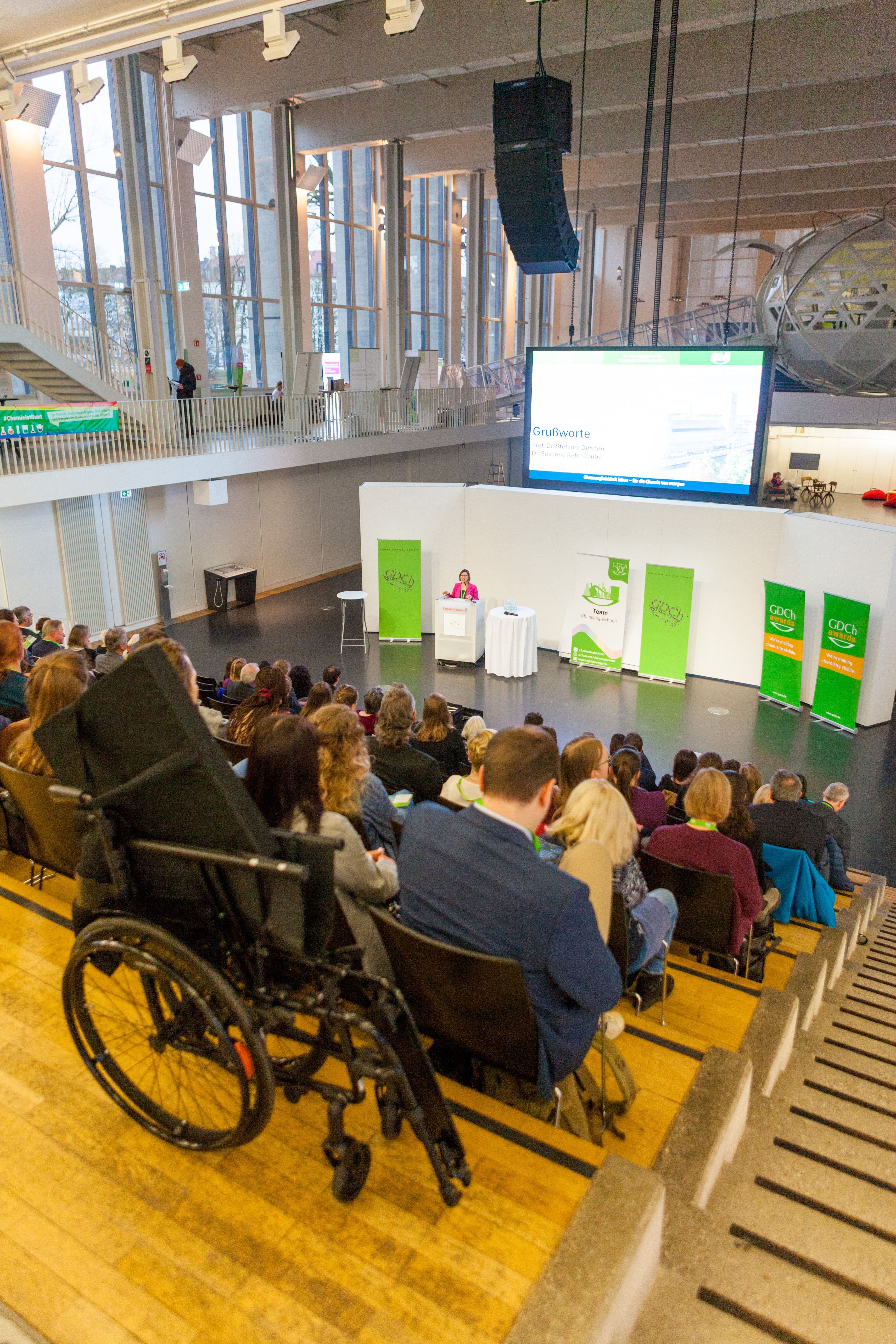 Blick auf eine Veranstaltung in einer modernen zweistöckigen Halle mit großen Glasfenstern. Eine Rednerin steht auf der Bühne vor grünen Roll-up-Bannern, dahinter ist ein großer Bildschirm mit 'Grußworte' zu sehen. Das Publikum sitzt in Reihen und hört zu. Im Vordergrund steht ein Rollstuhl, der die Barrierefreiheit der Veranstaltung verdeutlicht. Die Architektur ist modern mit viel Glas und einer Galerie im oberen Stockwerk.