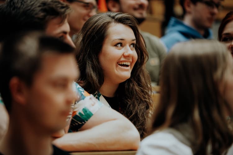 Mehrere Personen sitzen dicht beieinander in einem Raum mit Holzbänken. Im Vordergrund ist eine Person mit langen, braunen Haaren und einem gemusterten Oberteil zu sehen. Die Atmosphäre wirkt ruhig und konzentriert.